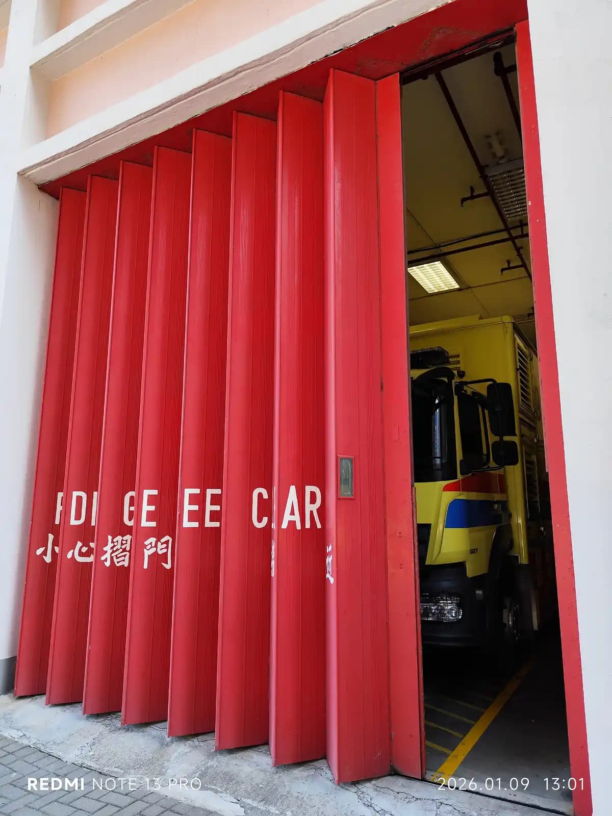 Porte pliante métallique partiellement repliée sur une baie de caserne, vantaux empilés en accordéon contre le piédroit