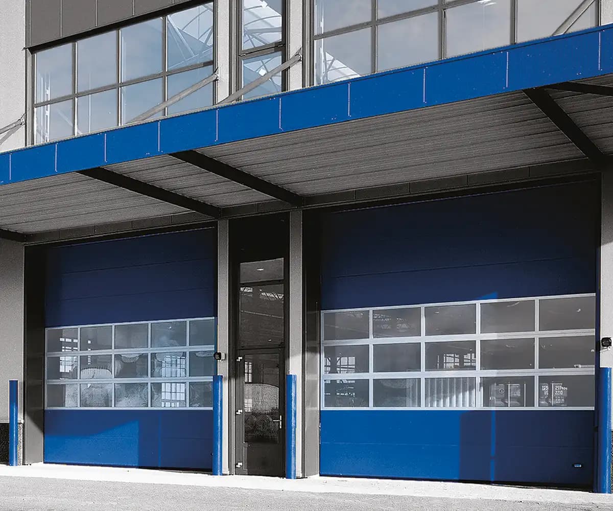 Deux portes sectionnelles industrielles bleues à hublots installées en façade d’un hall de production, sous auvent acier