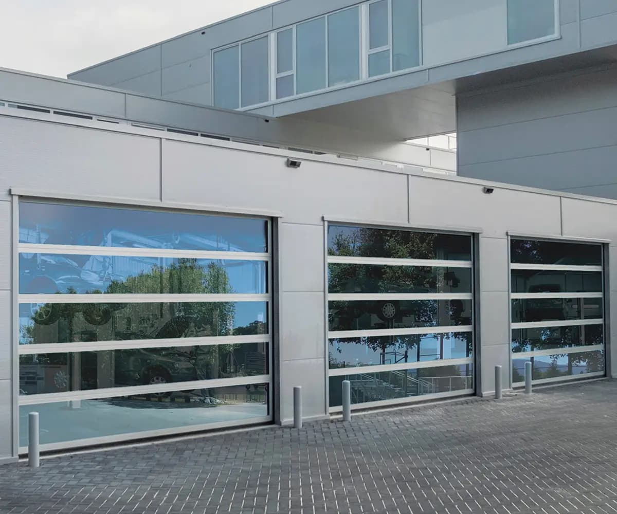 Variante entièrement vitrée : trois portes sectionnelles à panneaux aluminium full-vision sur une façade de concession automobile