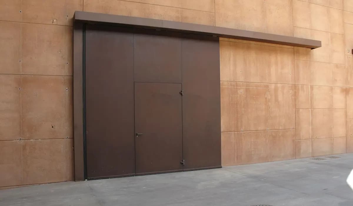 Porte coulissante industrielle à deux panneaux acier, ouverte et rangée à plat contre la façade sous son capotage de rail