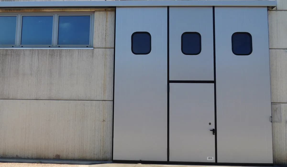 Vantail de porte coulissante en panneau isolé gris avec oculus carrés et portillon de service intégré
