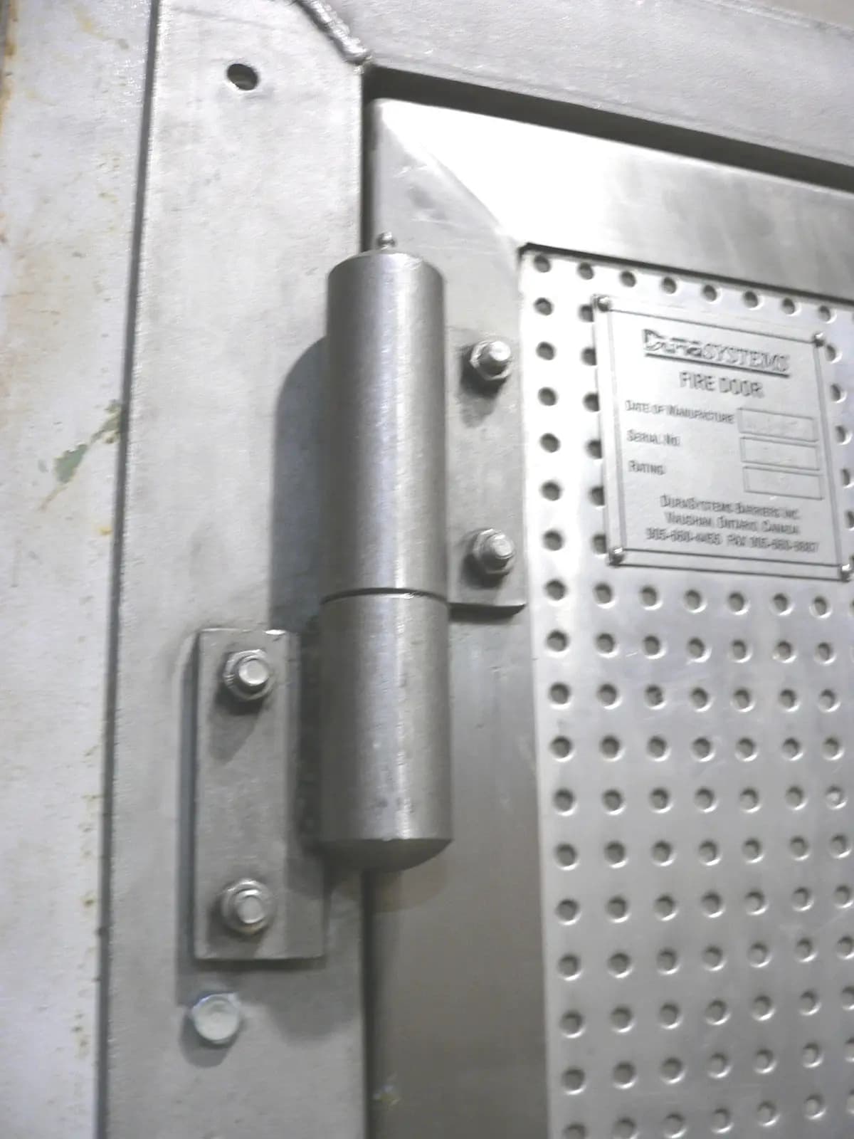 Paumelle acier lourde boulonnée sur un vantail coupe-feu inox, à côté de la plaque gravée d’identification de la porte