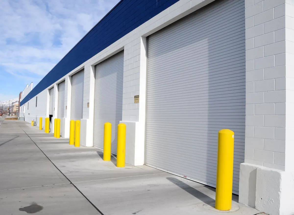 Rangée de portes enroulables industrielles en acier galvanisé fermées le long d’un bâtiment logistique, avec chasse-roues de protection