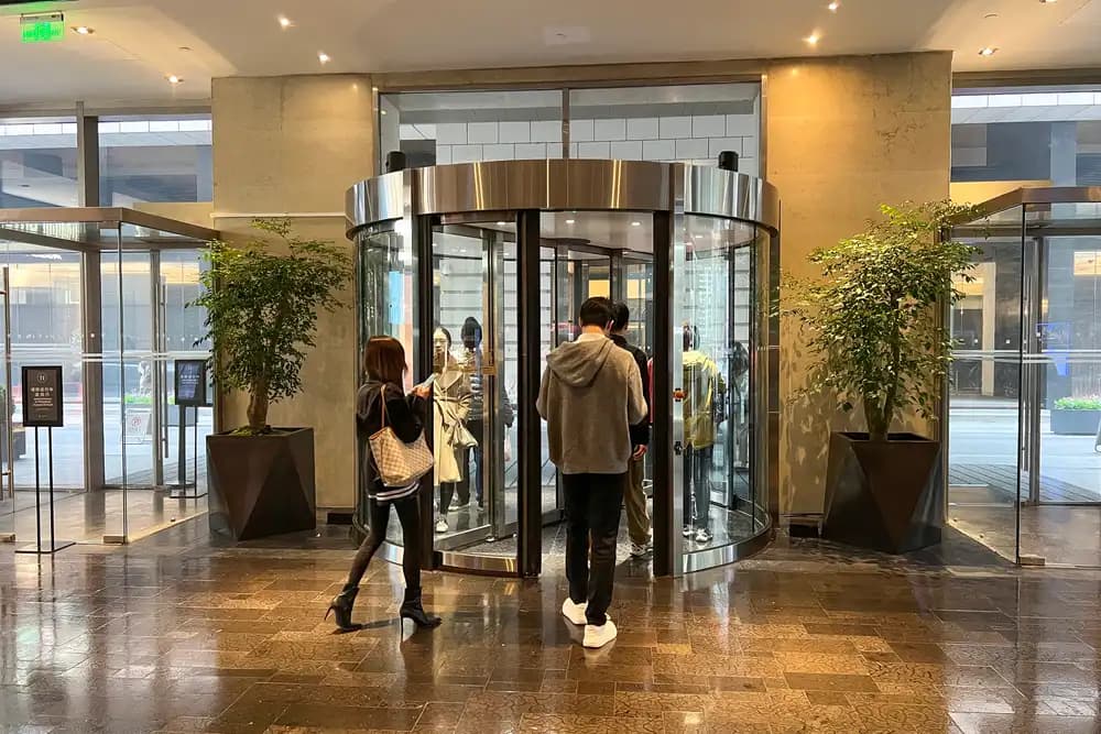 Porte tournante à bandeau inox en service dans un lobby d’hôtel en marbre, deux usagers la franchissant
