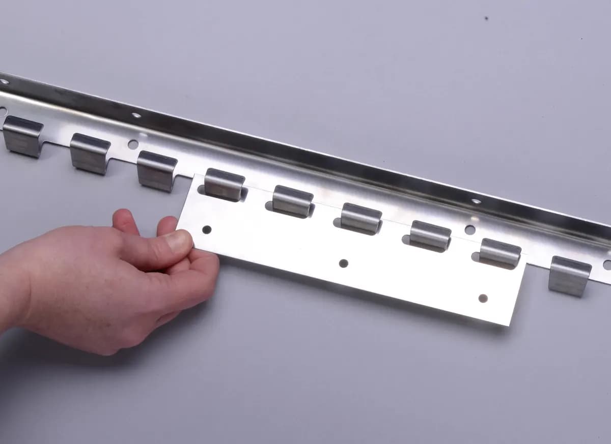 Détail de fixation : plaque à crochets inox présentée devant le rail de suspension perforé